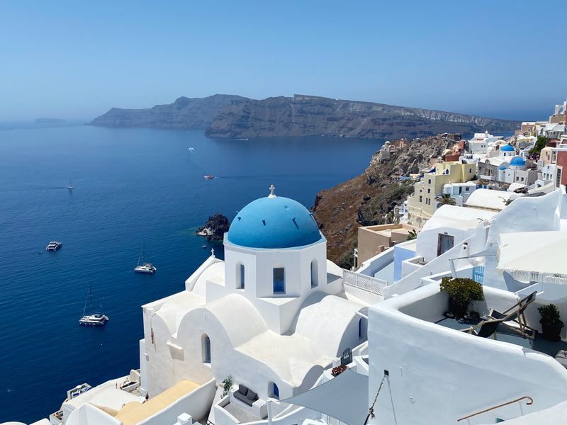 Weiße Kirche mit blauer Kuppel auf Santorini, Meer und Boote im Hintergrund.