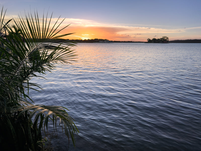 Sonnenuntergang über dem See von Flores mit ruhigem Wasser, warmem Himmel und Palmen im Vordergrund.